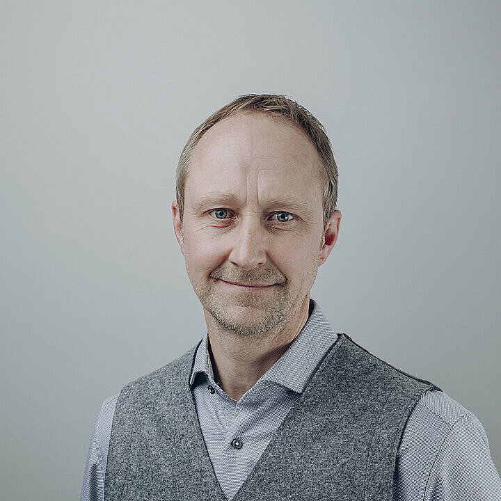 Xaver Wagner schmunzelnd in grauer Kleidung vor hellem Hintergrund.