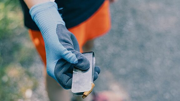 Hand mit blauer Arbeitshandschuh hält eine Metallzange mit einem kleinen Müllstück