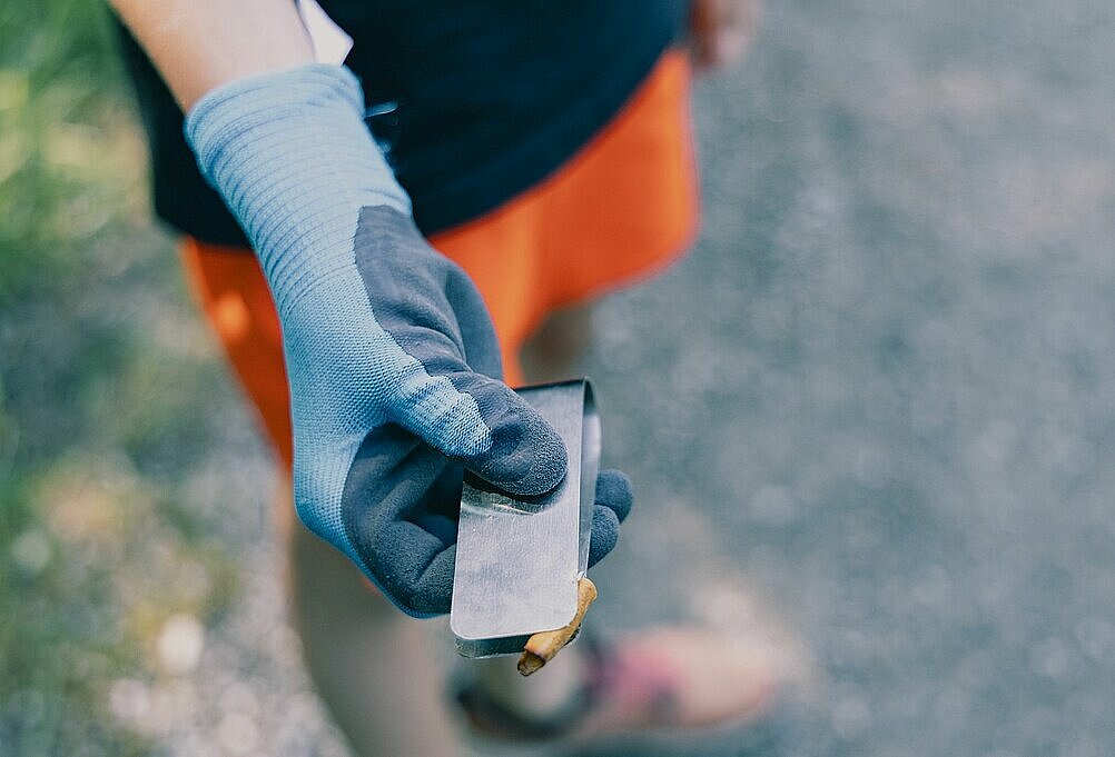 Hand mit blauer Arbeitshandschuh hält eine Metallzange mit einem kleinen Müllstück