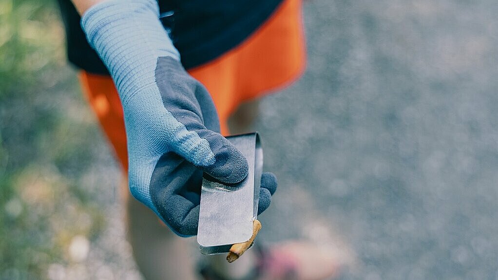 Hand mit blauer Arbeitshandschuh hält eine Metallzange mit einem kleinen Müllstück