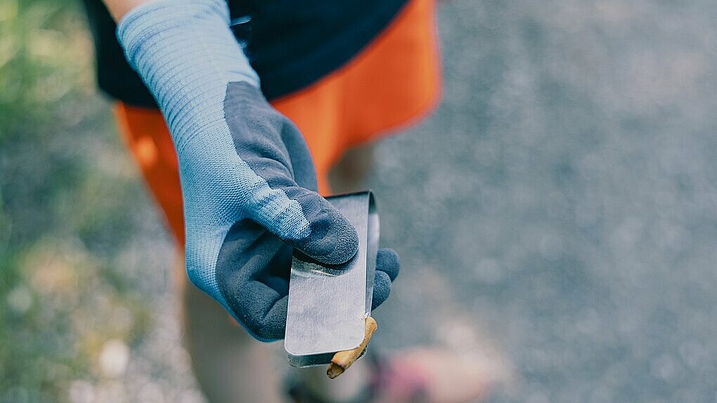 Hand mit blauer Arbeitshandschuh hält eine Metallzange mit einem kleinen Müllstück