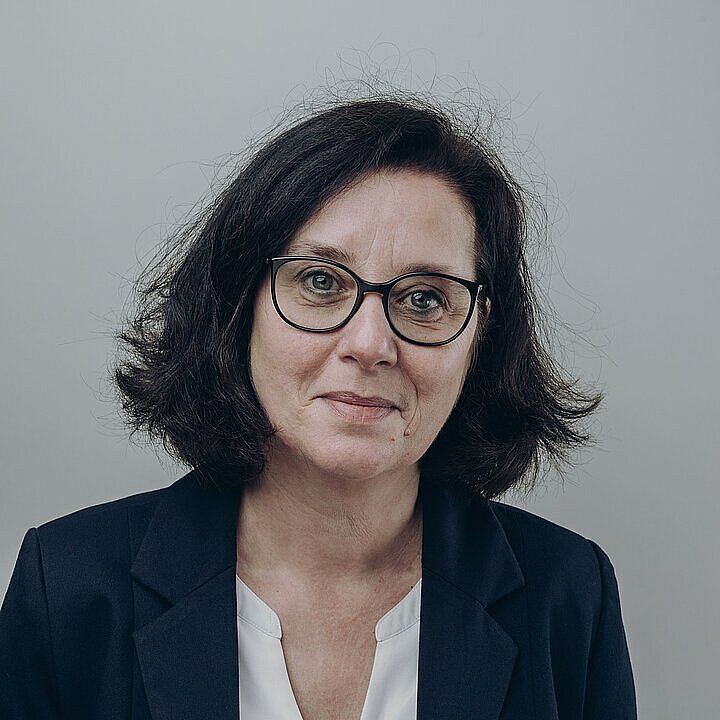 Claudia Mayer schmunzelnd mit weißer Bluse und schwarzem Blazer vor hellem Hintergrund.
