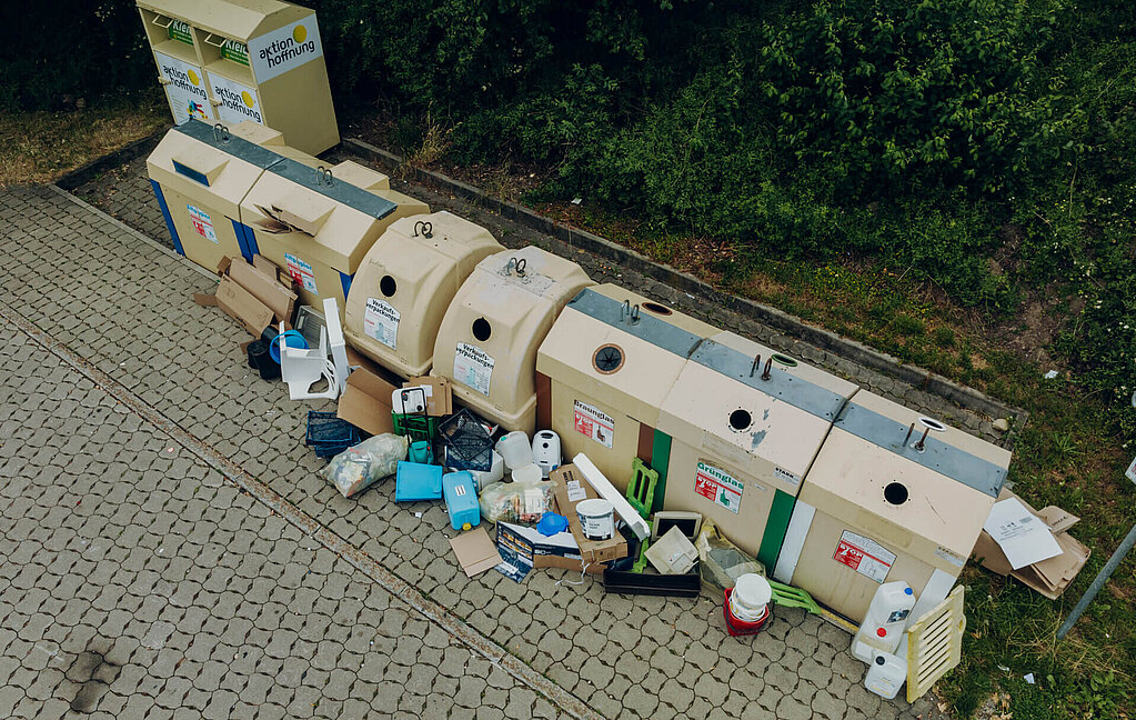 Wertstoffinsel mit mehreren Müllcontainern und danebenliegendem Abfall sowie Kartons auf gepflastertem Boden vor einem Grünstreifen