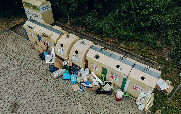 Wertstoffinsel mit mehreren Müllcontainern und danebenliegendem Abfall sowie Kartons auf gepflastertem Boden vor einem Grünstreifen